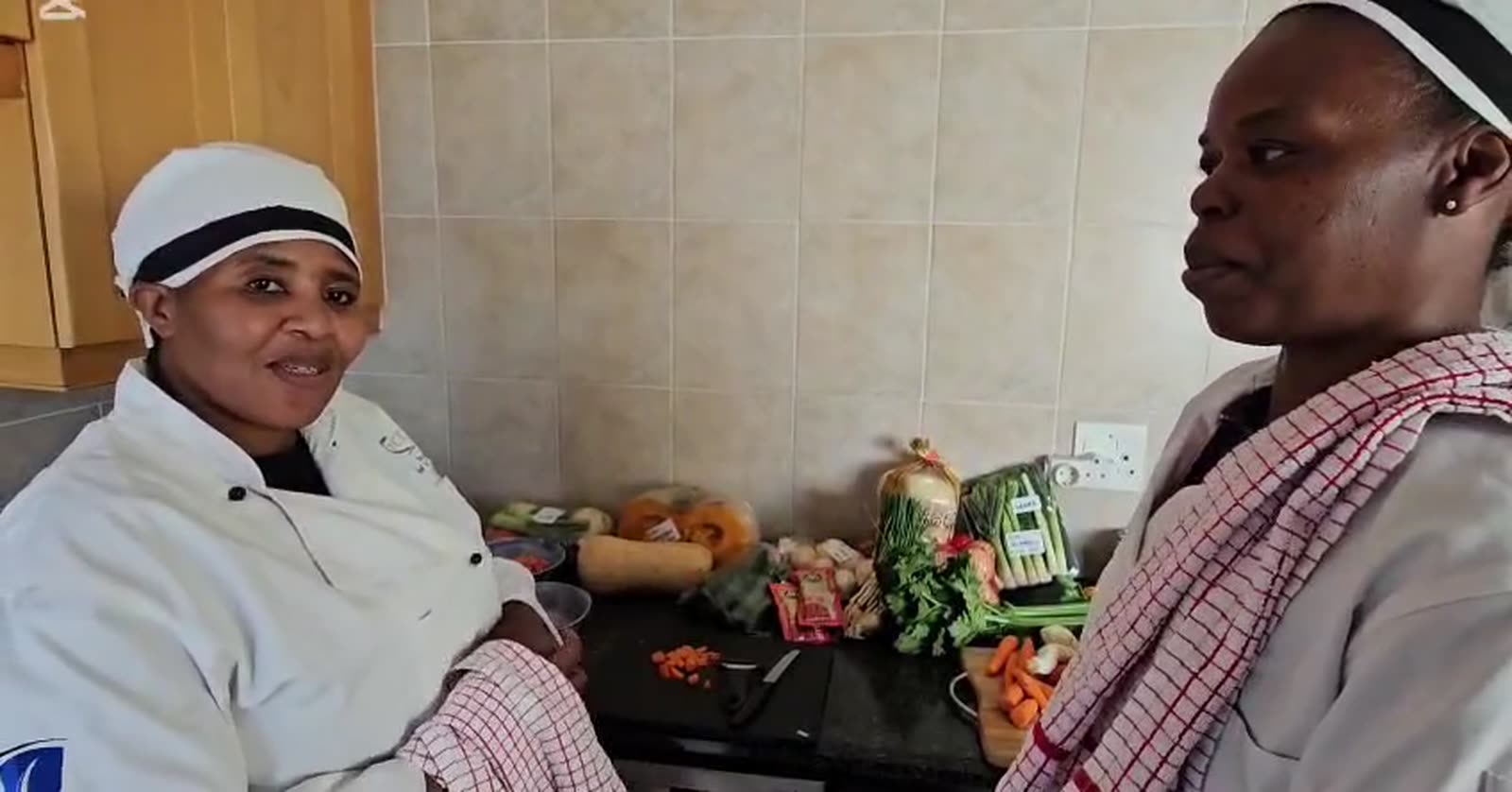 Cleo and Joyce in chef whites in their kitchen, fresh produce laid out on the counter